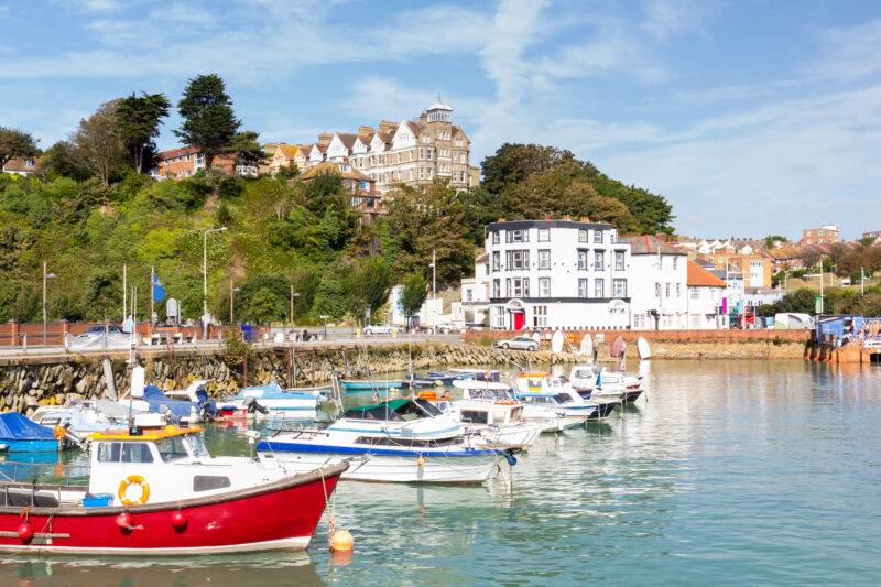 Picturesque harbor with small boats docked in the water, against a lush hillside with a mix of residential buildings and a prominent white building with a red door. The clear, sunny sky adds to the serene coastal setting.