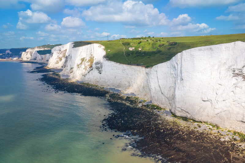 Tall white chalk cliffs with a grassy top, extending along a rocky shore. The sky is partly cloudy, enhancing the scenic beauty.