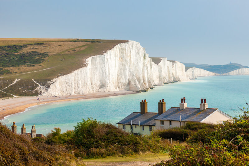 View of the white cliffs called the Seven Sisters near Eastbourne, with a wide blue bay in front and the grey slate roofs of two small cottages in the foreground.