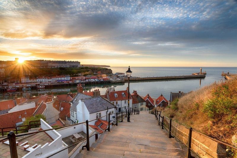 Looking fown a long stone staircase with several whitewashed cottages on the left and the sea at the bottom, it is sunset and the sun is just disappearing behind the harbour wall in Whitby North Yorkshire England