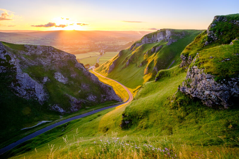 A winding road meanders through a lush green valley in the Peak District, flanked by steep hills, under a sunset sky. The golden light bathes the landscape,