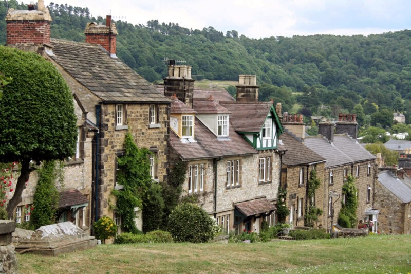 A row of traditional stone houses on a sloping hill in the village of Bakewell, with lush greenery and trees in the background. The houses are covered in ivy, have chimneys on the rooftops, and are closely packed together. The background features a dense forest.