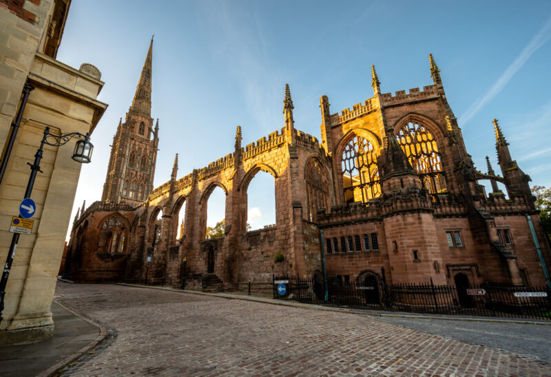 The ruins of Coventry Cathedral in Coventry, England, featuring tall Gothic arches and a prominent spire. The structure is bathed in the warm light of sunrise or sunset, highlighting the intricate stonework. Cobblestone streets and street lamps add to the historic ambiance of the scene.