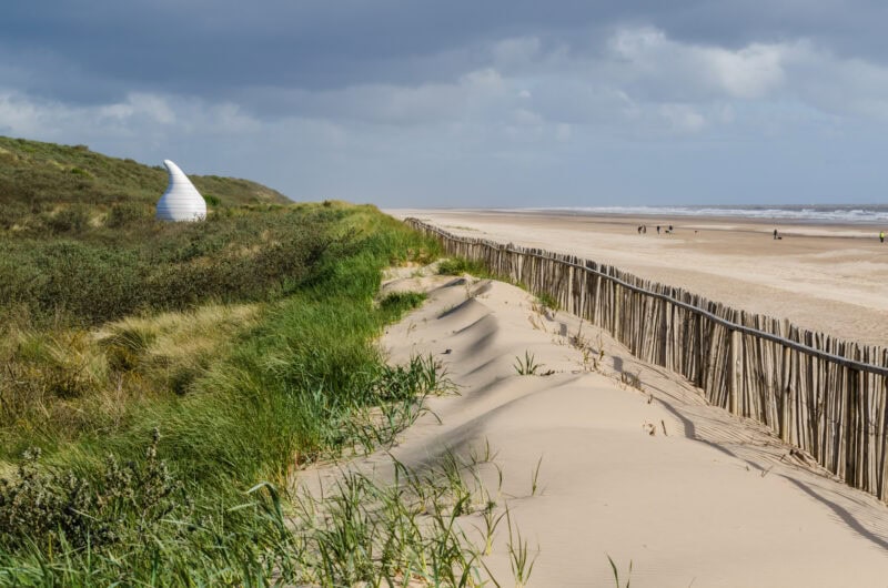 A coastal scene with a sandy beach and dunes. A wooden fence runs parallel to the beach, stabilizing the sand dunes. The dunes are covered with grass and vegetation. In the background, a white, spiral-shaped structure resembling a seashell is situated on a grassy hill. The beach stretches into the distance with a few people walking along the shoreline. The sky is cloudy, suggesting overcast weather.