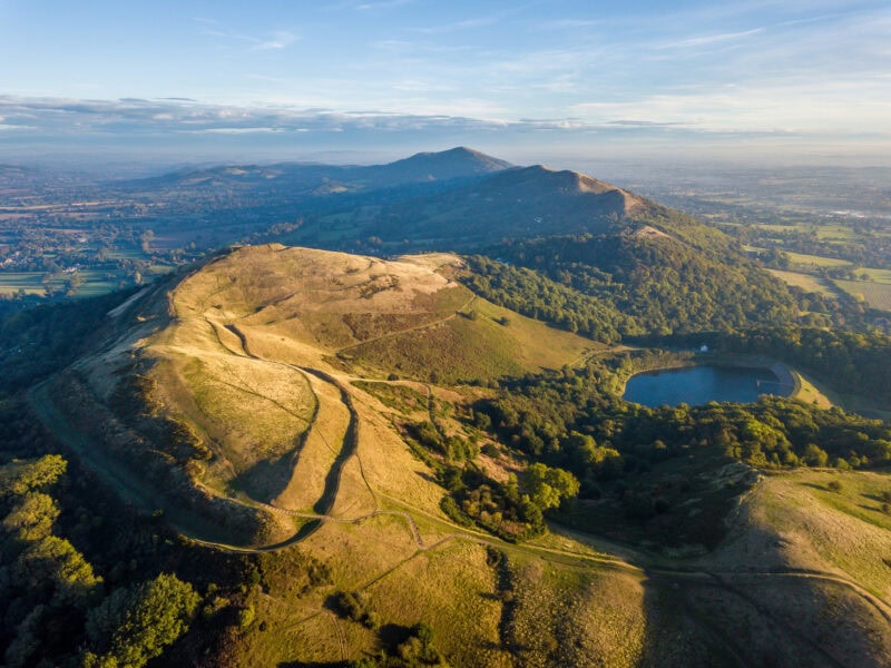 An aerial view of a hilly landscape in the Malvern Hills with a mix of grassy and wooded areas. Winding paths and trails traverse the hills, with a small lake or pond visible to the right. The sun casts long shadows, highlighting the terrain's contours and creating a picturesque scene of natural beauty