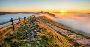 A scenic mountain ridge at sunrise with a stone path and wooden fence leading into the distance. The sun casts a warm glow over the landscape, with mist and clouds filling the valleys below. Mam Tor int he Peak District - Best Places to Visit in the Midlands England