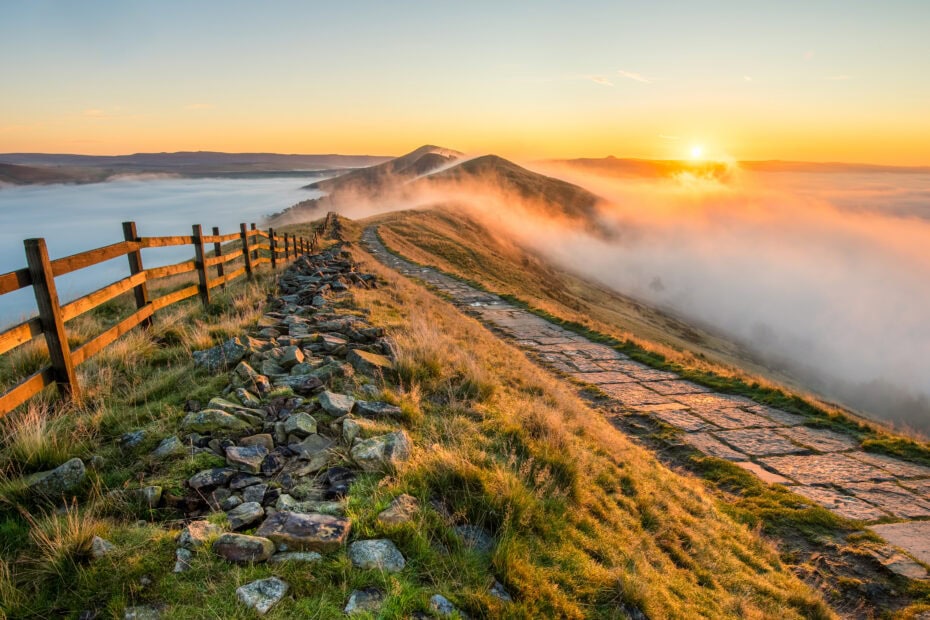 A scenic mountain ridge at sunrise with a stone path and wooden fence leading into the distance. The sun casts a warm glow over the landscape, with mist and clouds filling the valleys below. Mam Tor int he Peak District - Best Places to Visit in the Midlands England