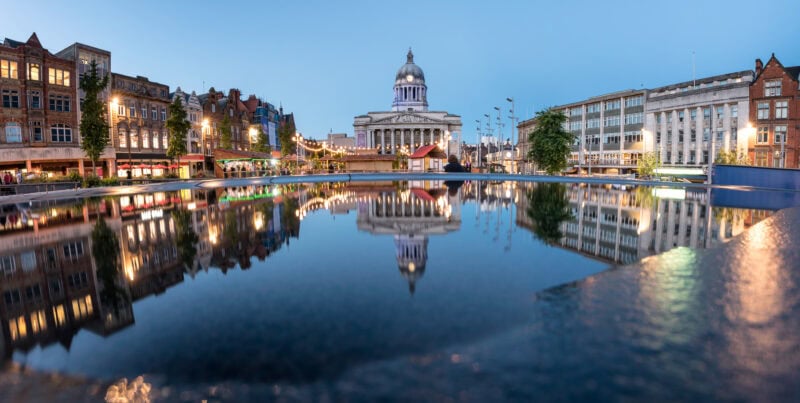 Panoramic view of the Old Market Square in Nottingham during twilight with the prominent domed building of City hall  at the center. Various buildings, including shops and offices, surround the square. Lights from the buildings reflect on a large, still body of water in the foreground, creating a symmetrical and visually appealing effect. The transitioning sky from day to night adds a serene quality to the scene.