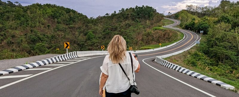 emily wearing denim shorts and a white short sleeved shirt walking away from the camera down the double yellow lines at the middle of a tarmac road which winds away over the hills into the distance | Tips to avoid being ronbbed while travelling