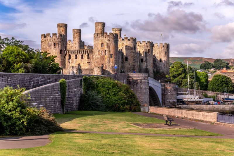 The historic Conwy Castle with multiple round towers and battlements, situated on a slight elevation. A stone bridge leads to the castle, surrounded by green trees and a well-maintained grassy area with a pathway and bench. 