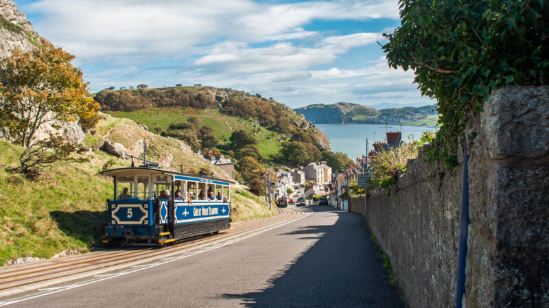 A tram labeled "5," travels along a track in a hilly area above Llandudno in North Wales. The tram is blue and white with the text "Great Orme Tramway" on its side.