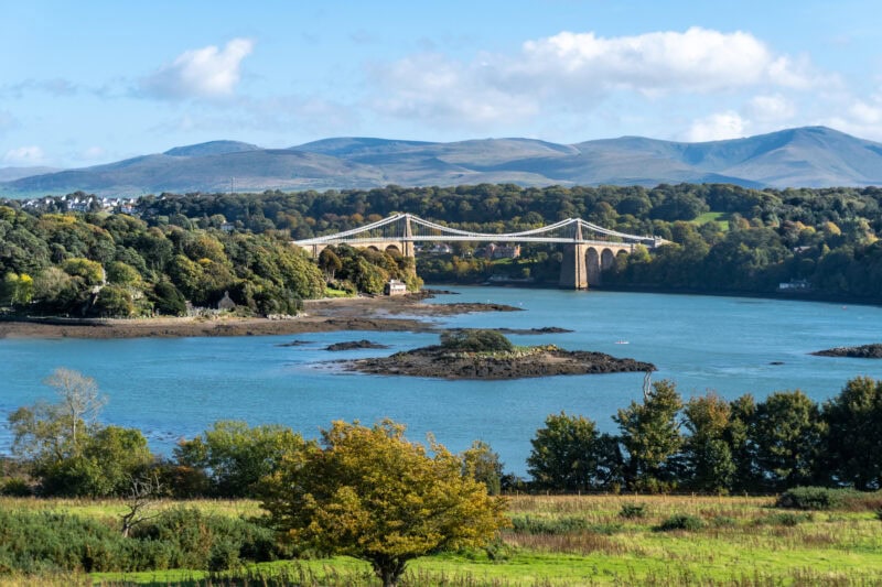 A suspension bridge spans a tranquil body of water in the Menai Strait, surrounded by lush green trees and foliage. Rolling hills and mountains form the backdrop under a partly cloudy sky. 