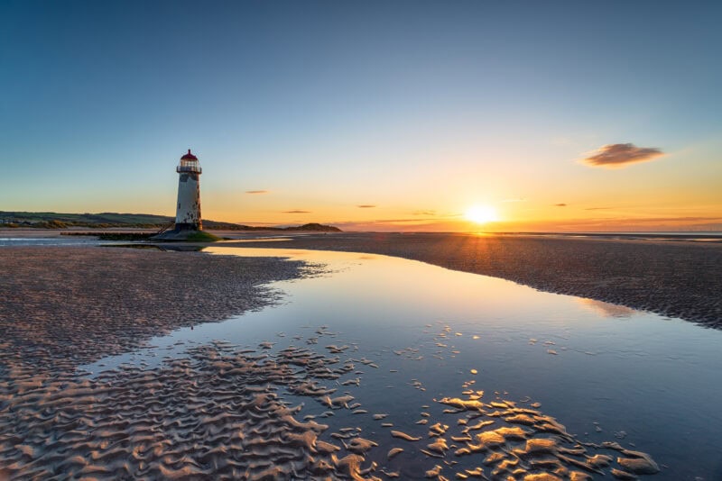 A lighthouse with a red top on a sandy beach at sunset. The beach features rippled sand patterns and a shallow pool of water reflecting the lighthouse and sky. 