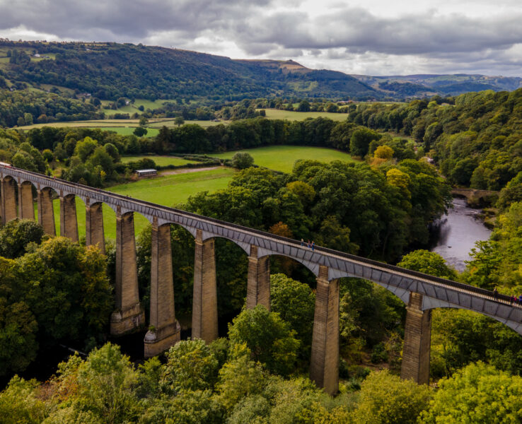 Aerial view of Pontcysyllte Aqueduct, a long, elevated stone structure spanning a lush, green valley with a river below. Supported by multiple tall, stone arches, it carries a narrow canal. 