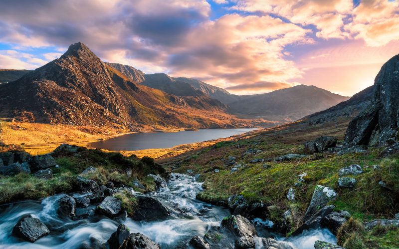 landscape in snowdonia national park with a stream in the foreground running towards a still lake at the foot of a mountain near sunset with pink clouds overhead in wales