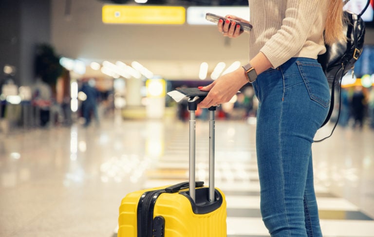 lower half of a woman in jeans holding a yellow suitcase and plane ticket in an airport - best carry on luggage for women