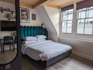 bed with gren velvet headboard and white duvet in a small serviced apartment in brixton london with wooden floor. there is a mirror in the foreground reflecting shelves and a tv on the opposite wall.