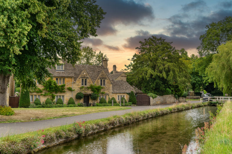 a shallow flat river with a row of beige stone cottages behind and several trees and a sunset sky behind - driving in the cotswolds england