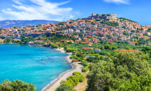 aerial view of Molyvos village in Lesvos with a curved bay and sandy beach next to turquoise water, the village is on the side of a hill beside the bay with a castle on top of the hill and red roofed buildings all around the side.