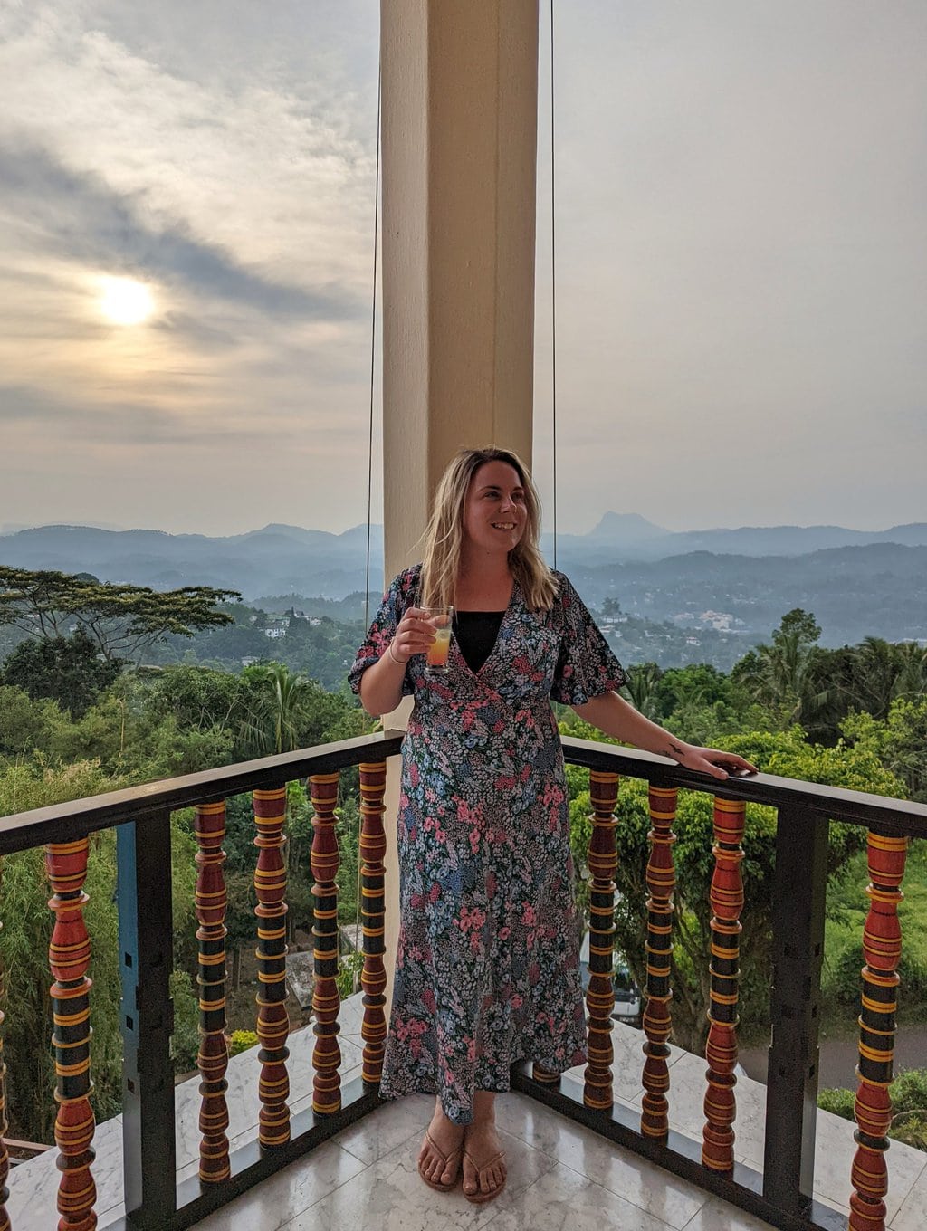 emily wearing a long dress with a blue and pink floral pattern with her blonde hair down and holding an orange drink in a small glass standing on a verandah with jungle and distand mountains in the background behind her just before sunset with a pale orange sky - how to make money from a travel blog