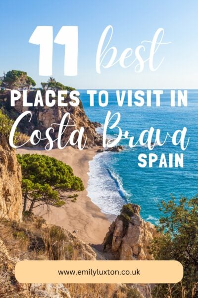 "11 Best Places to Visit in Costa Brava Spain" written in white text over a photo of a sandy beach surrounded by rocky cliffs topped with trees. The sea is very blue and it is a sunny day with clear sky. 