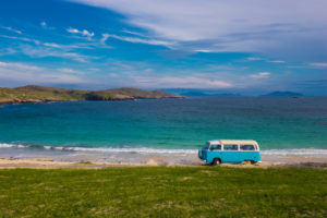 blue VW campervan driving on a road next to the sea in soctalnd with some low green cliffs in the distance behind on a very sunny day with blue sky above. best places to go in a campervan UK.