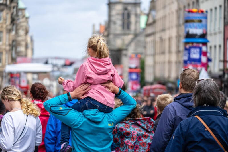 A crowd of people walking down a bustling street in Edinburgh Old Town, with a child in a pink jacket on an adult's shoulders.