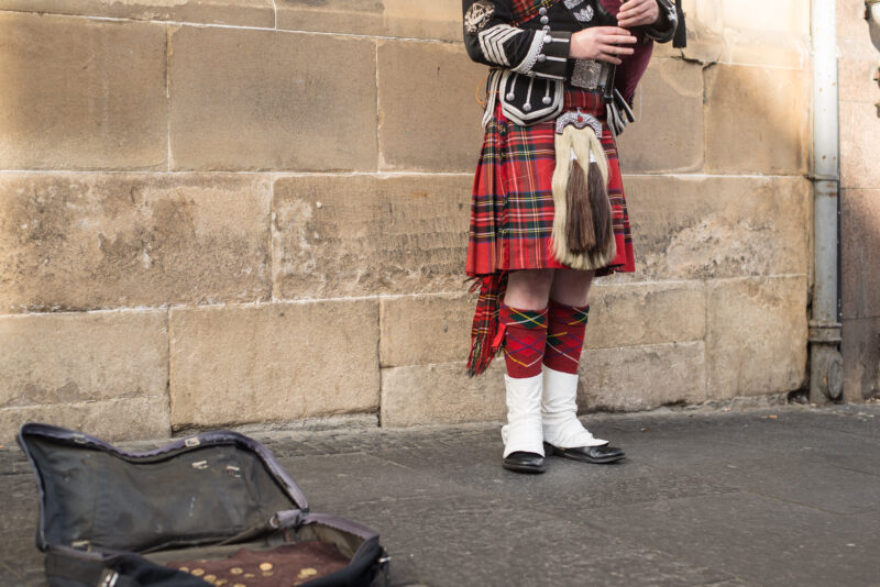 A person seen from the torso down wearing traditional Scottish attire, including a red tartan kilt, sporran, and red argyle socks, is playing a bagpipe in front of a stone wall. An open case with some coins is on the ground.