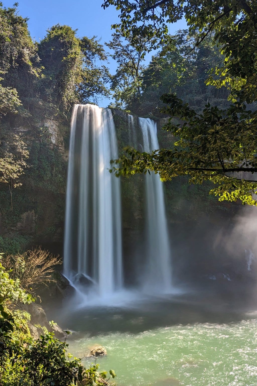 a tall waterfall taken with slow shutter so the water movement is blurred. the waterfall is emptying into a green pool surrounded by rocky cliffs and leafy trees.