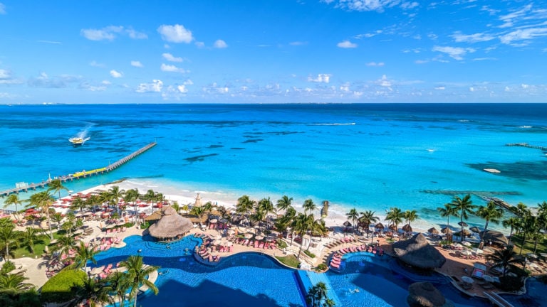 view from above of a large outdoor pool in frotn of a white sandy beach with the turquoise sea behind on a very sunny day. there is a narrow wooden pier to the left and a yellow board approaching. Grand Fiesta Americana Coral Beach Cancun review.