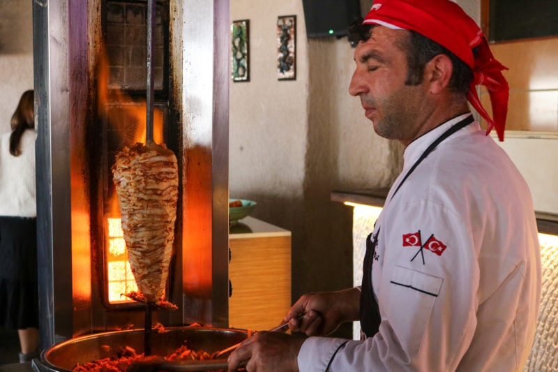 turkish man wearing a white shirt and red bandana using tongs to pick up meat from the metal tray in front of a large doner kebab on a spit in front of a flame