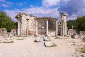 ruins of a curved brick wall with a cross carved into it and a row of 4 slim white stone pillars in front and the remains of a thicker white pillar to the right hand side on a sunny day with blue sky above