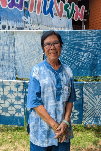 older thai lady with short dark hair wearing a blue and white tie-dyed kaftan top standing in front of several blue and white tie dyed fabrics and scarves hanging up in two layers behind her. Mo hom indigo dye in phrae. 