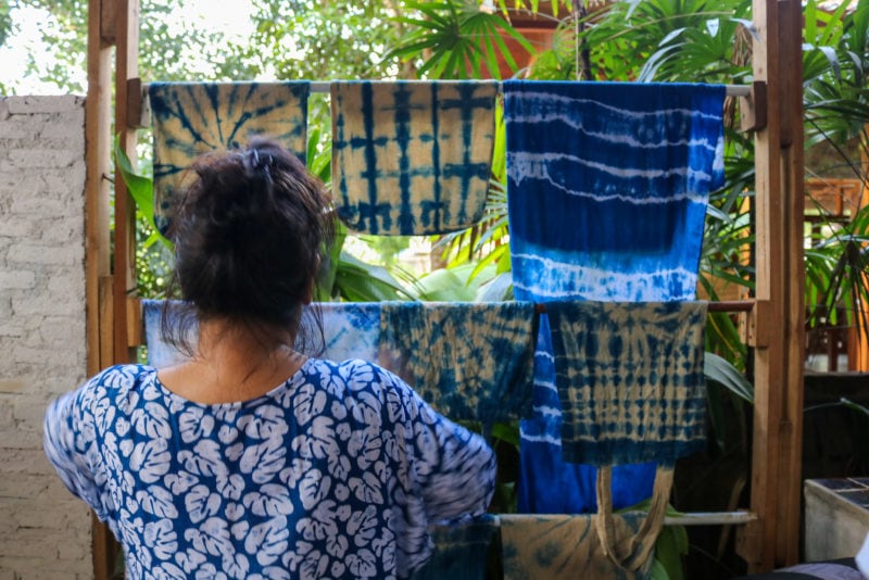 back of a thai woman with dark hair wearing a blue and white flowery patterned long sleeved top hanging something onto a double layered washing line with several indigo dyed clothes on it. things to do in phrae thailand.