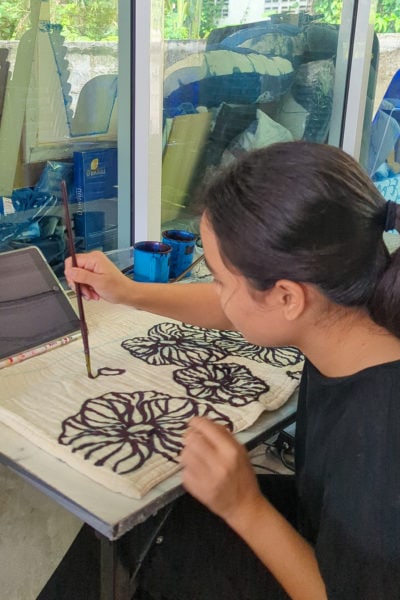 looking over the shoulder of a thai girl as she is painting large blue flowers onto a piece of white canvas using indigo dye