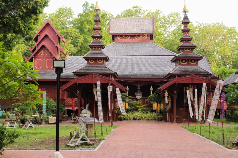 exterior of a large thai temple built from reddish coloured teak wood with a grey tiled roof and two multi-tiered pagodas in front on either side of the front door