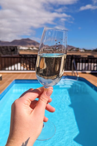 hand holding a glass of cava in front of an outdoor swimming pool with a view of volcanoes beyond in Lanzarote. Playa Blanca Holiday Villas review.