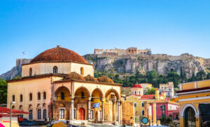 Large Ottoman mosque in Monastiraki square with yellow walls and a red tirled dowed roof, there are collonaded arches around the front section on the ground floor. Behind the church is a small rocky hill with the ancient ruins of the acropolis on top. unusual things to do in athens.