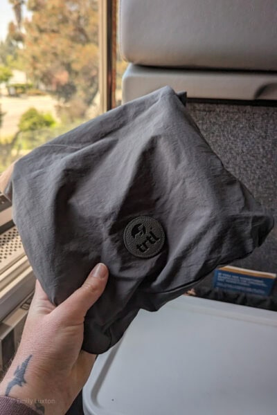 mão segurando uma pequena sacola de tecido cinza com um logotipo circular que diz TRTL em frente a uma janela de trem com árvores borradas do lado de fora