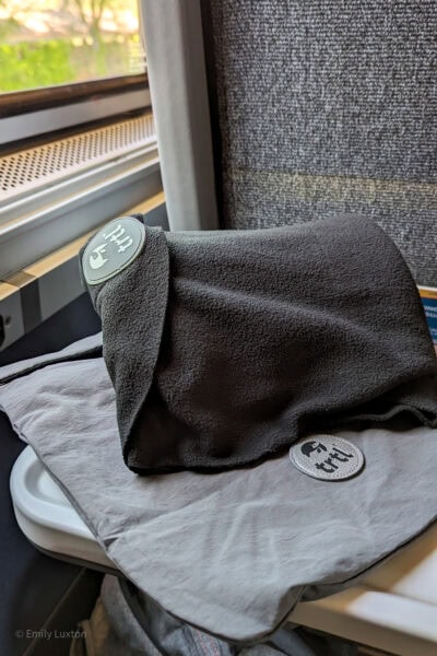 folded square of black fleece material with a grey logo saying TRTL. The fleece is on top of a grey bag with the same logo on a fold down tray table on a train with blurry trees visible out the window
