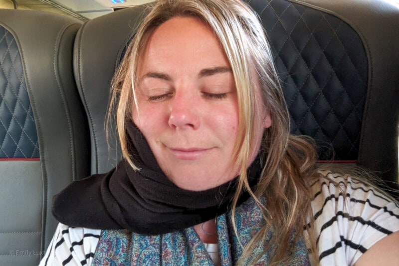 Selfie de Emily com os olhos fechados em um assento de trem de couro azul vestindo uma camiseta branca com listras pretas e um lenço de lã preto enrolado no pescoço