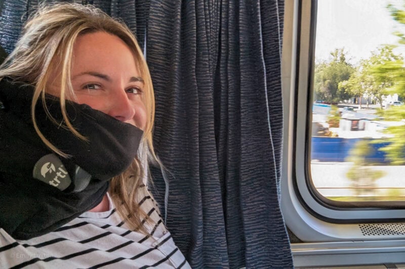 Selfie of Emily on a blue leather train seat wearing a white t shirt with black stripes and a black fleece scarf wrapped around her neck - TRTL Travel Pillow Review