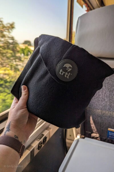 mão segurando o travesseiro de viagem TRTL em frente a uma janela de trem com árvores e céu azul do lado de fora, o travesseiro é um quadrado de lã dobrado com um círculo cinza contendo o logotipo TRTL.