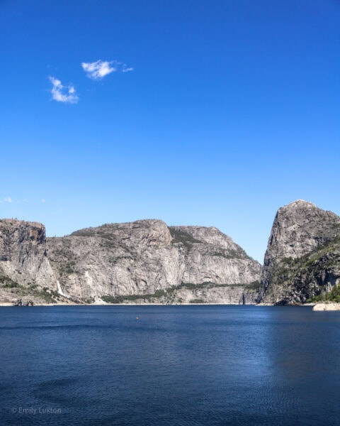 grande lago azul com picos rochosos cinzentos do outro lado e céu azul claro acima.
