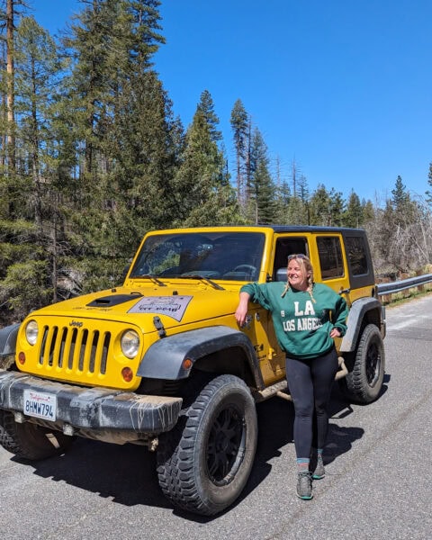 Emily standing leaning on a bright yellow jeep with pine forest behind and clewar blue sky above. She is wearing black leggings and a dark green jumper and has her hair in plaits.
