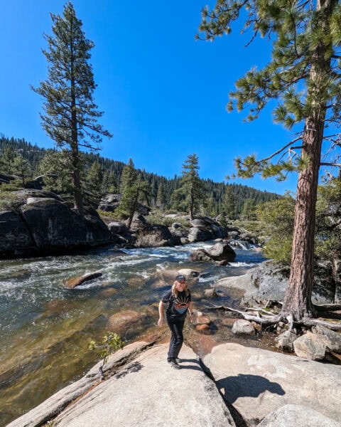 emily standing in front of rushing river with grey rocsk and pine trees on either side and a bright blue lake beyond with a hill covered in pine forest on the far side. she is wearing black trousers and a grey t shirt and her hair is in bunches.