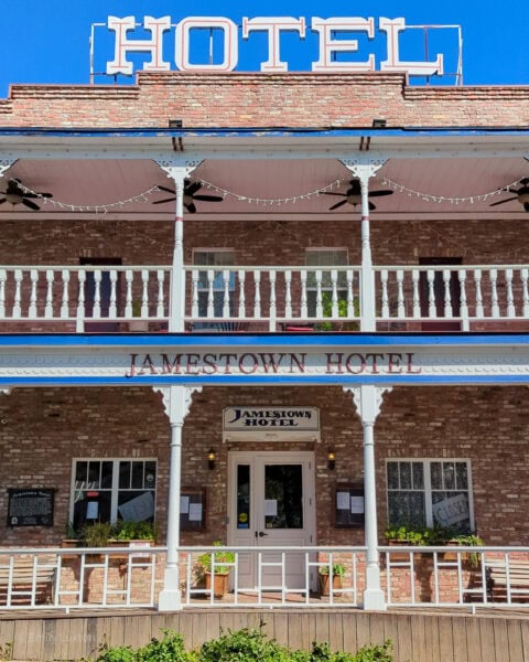 closeup of the front of the Jamestown Hotel, a brick building with a verandah on the ground floor and a wooden balcony on the top floor. Large white letters saying HOTEL are on the roof.
