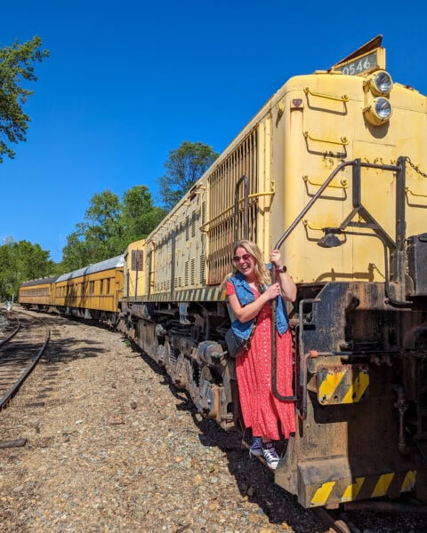 Emily sanding on the steps of a yellow Diesel engine leaning on one of the rails. Emily is wearing a long red dress and a blue denim waistcoat and has her long hair loose. Taken at Railtown State Historic Park in Tuolumne County California.