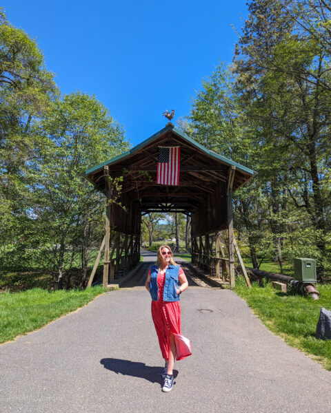 Emily standing with her hands in her pockets in front of a wooden covered bridge with a US flag hanging from the top of it and trees on either side. Emily is wearing a long red dress and a blue denim waistcoat and has her long hair loose. The entrance to Indigeny Reserve in Tuolumne County California.