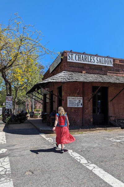 emily walking across a road in front of a red brick single storey building with a white sign that says St Cjharles Saloon. Emily is wearing a long red dress and a blue denim waistcoat and has her long hair loose.